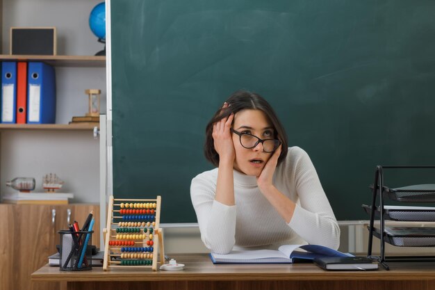 surpris de mettre les mains sur le visage jeune enseignante assise au bureau avec des outils scolaires en classe