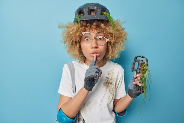 Surpris, une mystérieuse femme bouclée fait un geste de silence dit des regards secrets embarrassés essaie de réparer son vélo après avoir roulé porte un équipement de protection t-shirt sale isolé sur fond bleu