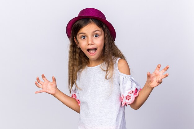 Surpris petite fille de race blanche avec chapeau de fête violet en gardant les mains ouvertes isolé sur un mur blanc avec espace copie