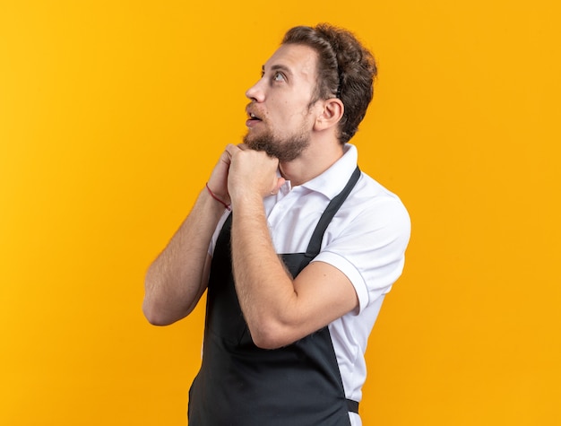 Surpris à la recherche d'un jeune coiffeur masculin en uniforme mettant les mains sous le menton isolé sur fond jaune