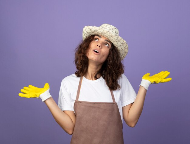 Surpris à la recherche d'une jeune femme jardinière en uniforme portant un chapeau de jardinage et des gants écartant les mains