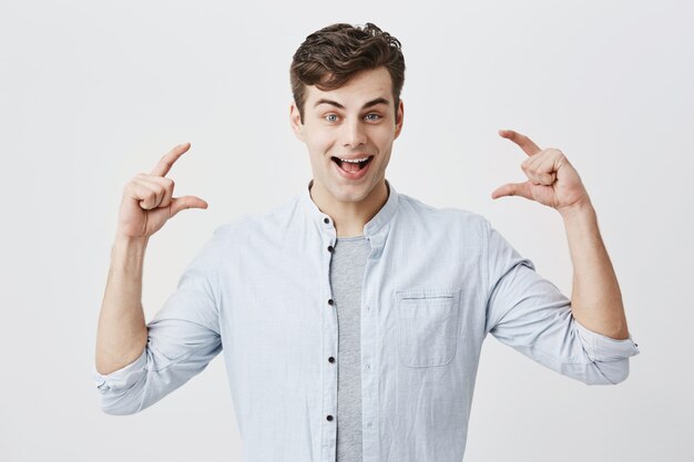 Surpris surpris jeune mannequin européen souriant joyeusement avec la bouche ouverte, démontrant la taille avec les deux mains de quelque chose de petite taille, faisant des gestes activement. Langage corporel et gestes.