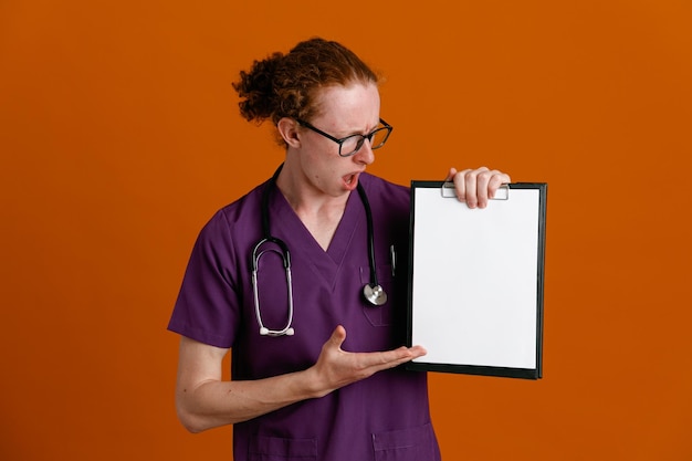 Surpris tenant et regardant le presse-papiers jeune médecin de sexe masculin portant l'uniforme avec stéthoscope isolé sur fond orange