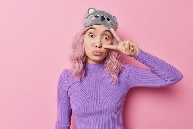 Surprise, une femme asiatique séduisante aux cheveux teints en rose fait un geste de paix sur les yeux, regarde des gestes émerveillés et montre un signe v porte un masque de sommeil et un col roulé violet isolé sur fond de studio rose