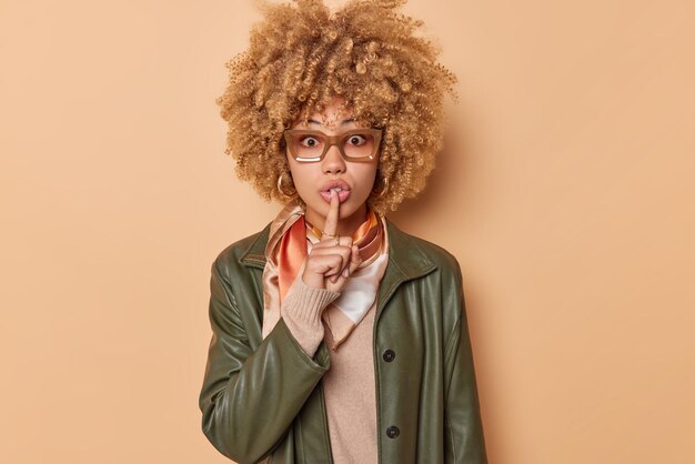 Surprise, une jeune femme aux cheveux bouclés se sent choquée fait un geste de silence chut à l'intérieur raconte des rumeurs porte des lunettes veste en cuir isolée sur fond marron. Ne dis mon secret à personne