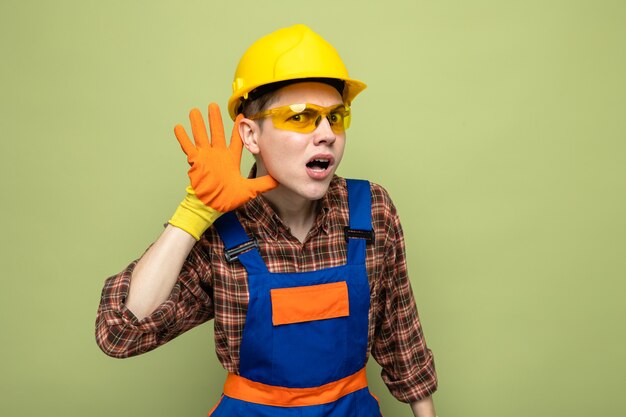 Suspect montrant le geste d'écoute jeune constructeur masculin portant l'uniforme et des gants avec des lunettes