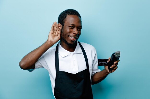 Suspicieux montrant écouter geste tenant la carte avec des tondeuses à cheveux jeune coiffeur afro-américain en uniforme isolé sur fond bleu
