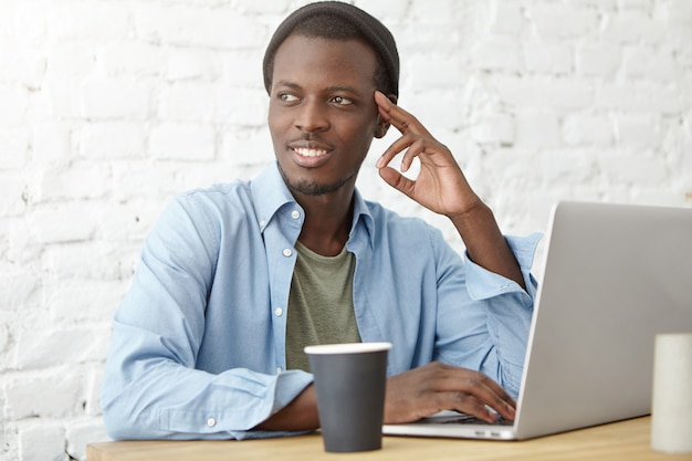 Photo gratuite sylish étudiant afro-américain prenant un café pendant le déjeuner à la cafétéria, utilisant un ordinateur portable tout en travaillant sur un projet de diplôme ou en préparant des cours. jeune hipster noir appréciant le petit déjeuner au café