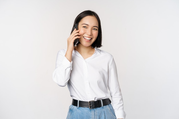 Sympathique femme asiatique souriante parlant au téléphone fille sur appel tenant un smartphone et riant parlant debout sur fond blanc