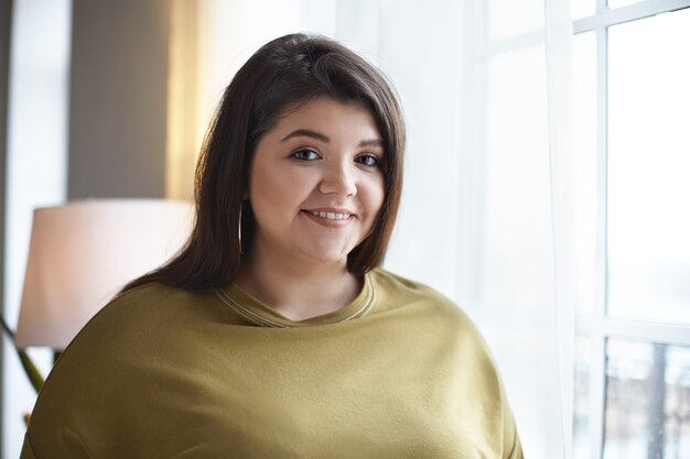 Sympathique à la jeune femme joyeuse avec un corps sinueux et des cheveux noirs lâches bénéficiant d'une belle vue urbaine tout en se tenant par la fenêtre à la lumière du jour, souriant joyeusement Concept de personnes et de style de vie