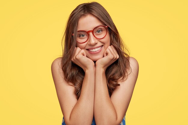 sympathique jeune femme avec des lunettes posant contre le mur jaune