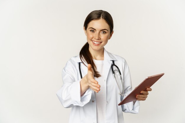 Sympathique jeune femme médecin, médecin tend la main pour la poignée de main, saluant le patient à la clinique, tenant une tablette numérique avec des données médicales, fond blanc.