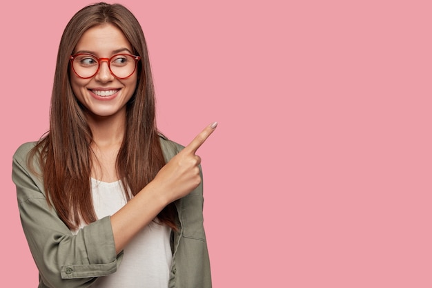 Sympathique jeune femme souriante aux cheveux bruns, donne de bons conseils ou suggère quoi acheter, indique avec l'index dans le coin supérieur droit