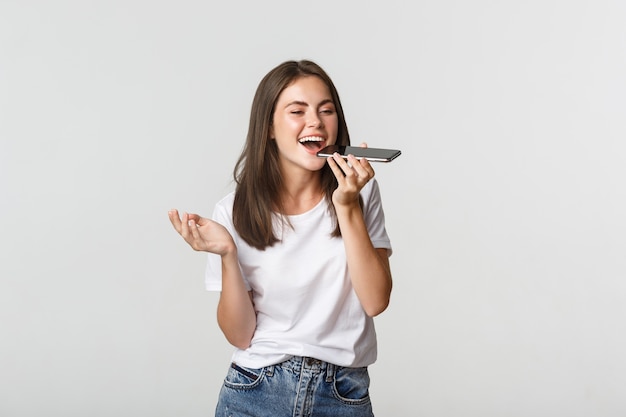 Sympathique jolie fille brune parler sur haut-parleur, enregistrer un message vocal sur smartphone, blanc.