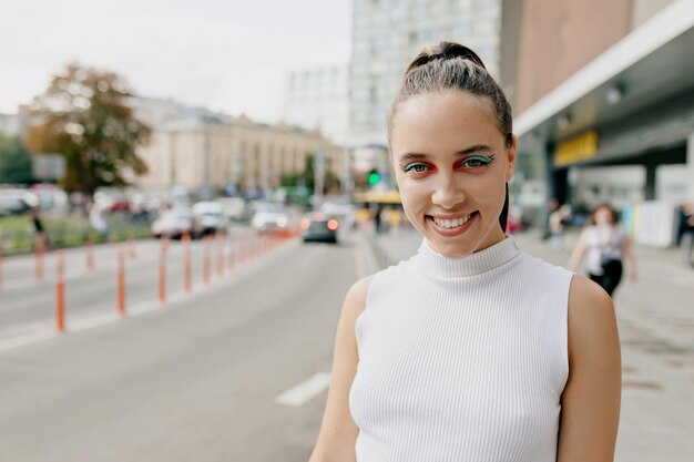 Sympathique jolie fille avec un maquillage brillant et des cheveux foncés habillés d'un t-shirt blanc sourit à la caméra et passe du temps à l'extérieur dans le centre-ville par une chaude journée ensoleillée