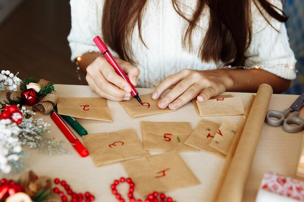 Une table avec des décorations de noël et les mains d'une fille écrivant des chiffres sur un calendrier de l'avent bricolage