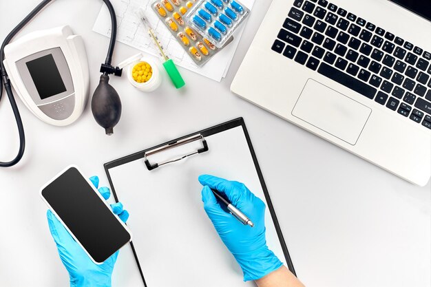 Table de médecin, outils, instruments médicaux, tonomètre de thérapeute, tension artérielle, travail à l'hôpital sur fond blanc à plat. La main du médecin dans un gant bleu enregistre les indicateurs sur un papier blanc