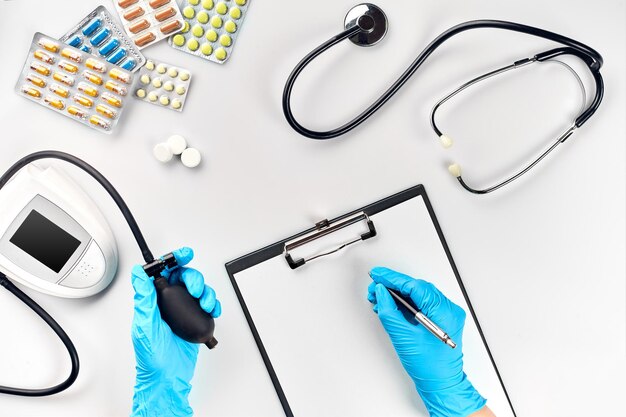 Table de médecin, outils, instruments médicaux, tonomètre de thérapeute, tension artérielle, travail à l'hôpital sur fond blanc à plat. La main du médecin dans un gant bleu enregistre les indicateurs sur un papier blanc