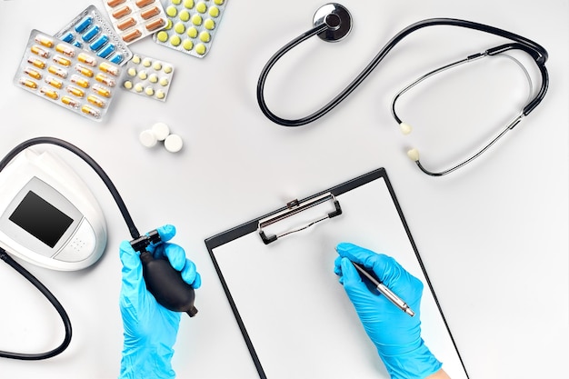 Table de médecin, outils, instruments médicaux, tonomètre de thérapeute, tension artérielle, travail à l'hôpital sur fond blanc à plat. La main du médecin dans un gant bleu enregistre les indicateurs sur un papier blanc