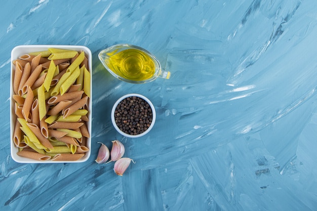 Un tableau blanc de pâtes crues avec de l'huile et des grains de poivre sur fond bleu.