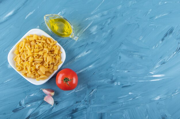 Un tableau blanc de pâtes crues avec de l'huile et des tomates rouges fraîches sur fond bleu.