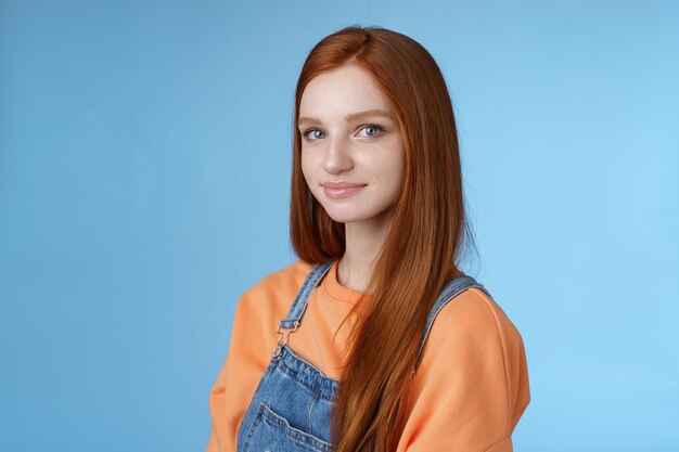 Taille gentille sincère tendre belle fille rousse vêtue d'une salopette en jean orange, debout à moitié tournée, souriante, stupide et souriante, caméra à la recherche amicale, marchant agréablement seule sur fond bleu.