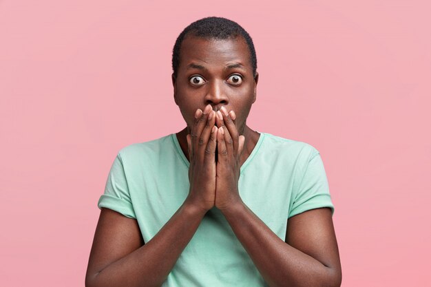 Taille portrait de beau jeune homme afro-américain regarde dans la terreur avec les yeux sortis, couvre la bouche, exprime un grand choc