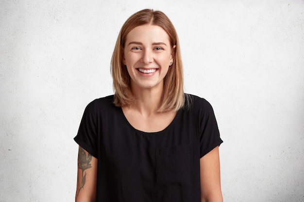 Taille portrait de la belle jeune femme souriante avec une expression positive, porte un t-shirt noir décontracté, a un bras tatoué, des modèles contre un mur de béton