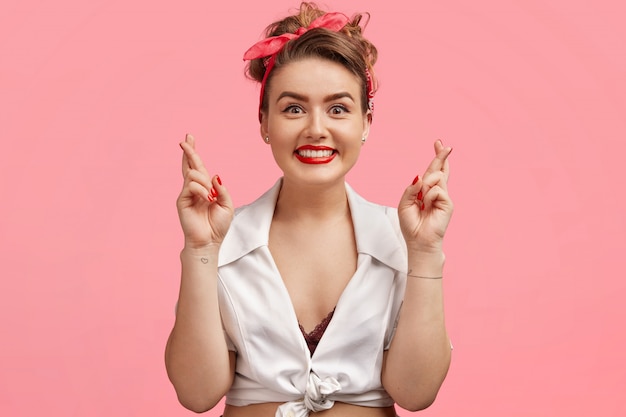 Taille portrait de pin-up beau modèle féminin avec des lèvres peintes en rouge et manucure, a un style rétro, croise les doigts avec une expression positive, espère quelque chose, a un but dans la vie