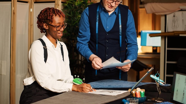 Photo gratuite tailleur et apprenti examinant des croquis en atelier, stagiaire apprenant le concept artisanal pour créer des vêtements. couturier expérimenté expliquant les techniques de couture aux débutants dans un atelier de tailleur.