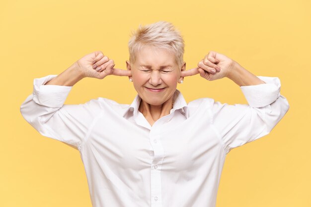 Tais-toi! Image isolée d'une femme mature en colère frustrée avec des cheveux de lutin teints gardant les yeux fermés et bouchant les oreilles, ne supportant pas les sons forts ou le bruit, étant stressée tout en se battant ou en se disputant