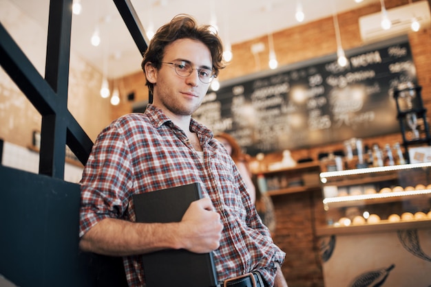 Talentueux jeune homme en chemise décontractée tenant un cahier dans un café.