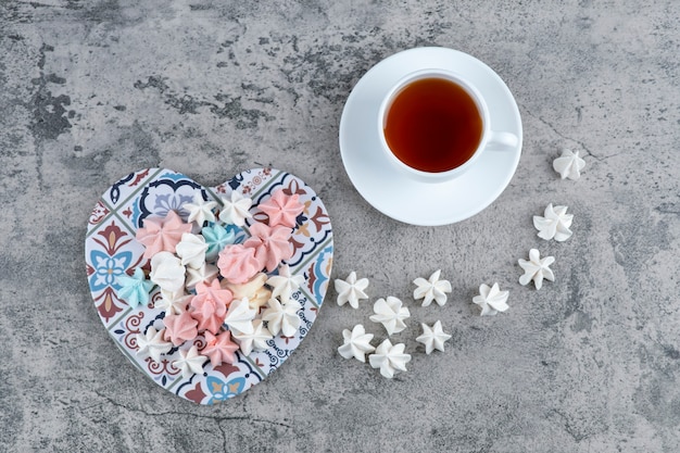 Tas de bonbons meringués colorés sur un dessous de plat en forme de coeur et une tasse de thé.