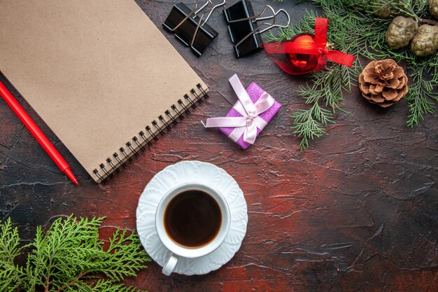 Une tasse d'accessoires de décoration de branches de sapin de thé noir et un cadeau à côté d'un cahier avec un stylo sur fond sombre