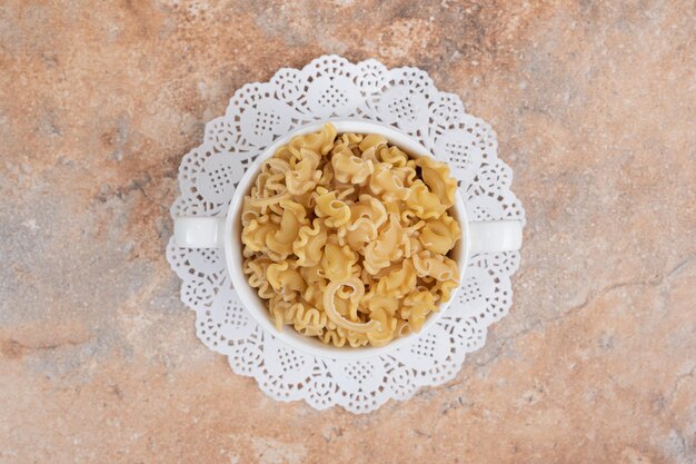 Une tasse blanche pleine de macaronis frais non préparés sur un espace en marbre.