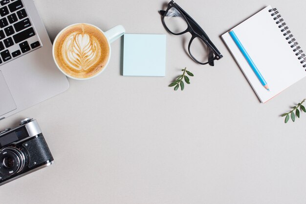 Tasse à café cappuccino; portable; appareil photo rétro; bloc-notes adhésif; lunettes et crayon sur le bloc-notes en spirale sur fond blanc