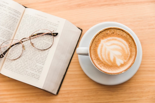 Tasse à café chaude avec de l&#39;art cappuccino latte et des lunettes sur un livre ouvert sur la table