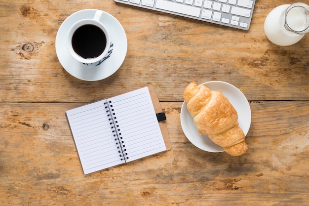 Photo gratuite tasse à café; croissant cuit au four; lait avec clavier et bloc-notes à spirale blanche sur un bureau en bois