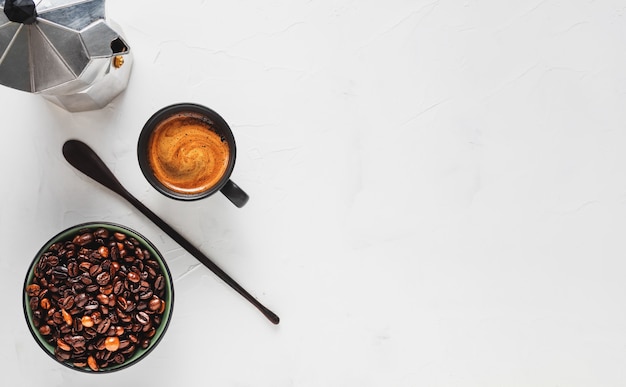 Tasse à café avec expresso fort avec mousse, une cafetière et des grains de café dans un bol sur une surface en béton blanc