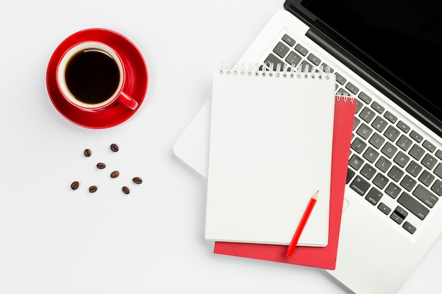 Tasse à café, grains de café avec le bloc-notes en spirale sur un ordinateur portable ouvert sur fond blanc