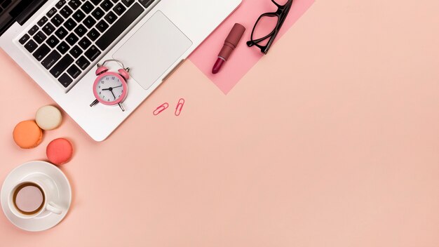 Tasse à café, macarons, réveil, ordinateur portable, lunettes et rouge à lèvres sur fond de pêche