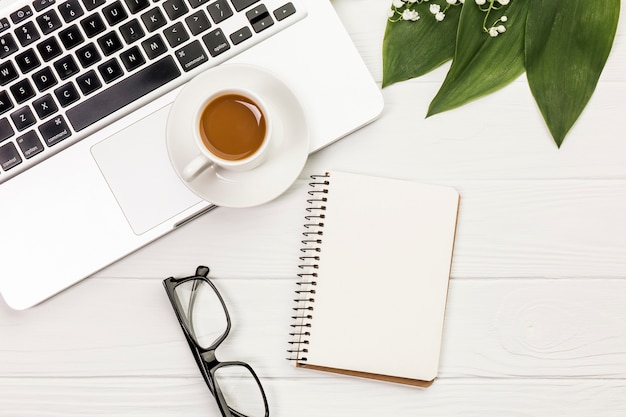 Tasse à café sur ordinateur portable, lunettes, bloc-notes à spirale et feuilles sur un bureau blanc