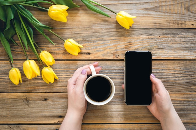 Une tasse de café et un smartphone dans des mains féminines et un bouquet de tulipes jaunes