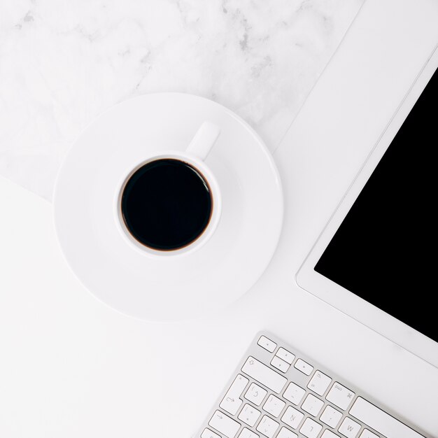 Tasse à café sur une soucoupe près de la tablette numérique et clavier sur un bureau blanc