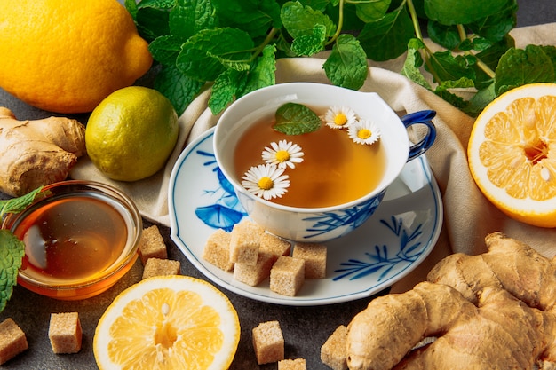 Tasse de thé à la camomille avec des citrons, du gingembre, des cubes de sucre brun, du miel dans un bol en verre et des feuilles vertes dans une soucoupe sur fond gris et morceau de tissu, gros plan.