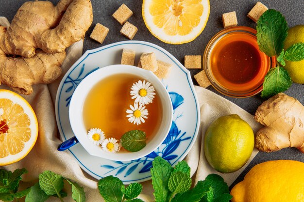 Tasse de thé à la camomille avec tranches de citron, gingembre, morceaux de sucre brun, miel dans un bol en verre et feuilles vertes dans une soucoupe sur fond gris et morceau de tissu, à plat.