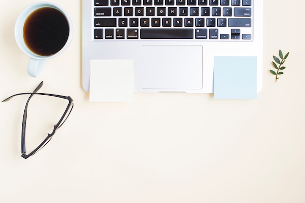 Tasse à thé; lunettes et bloc-notes adhésif blanc sur un ordinateur portable sur fond beige