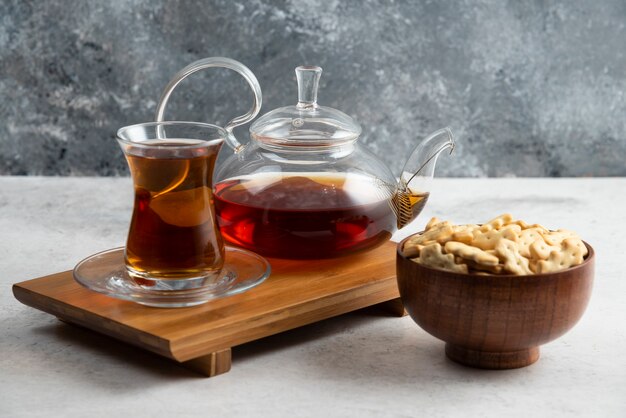 Une tasse de thé en verre avec un bol en bois plein de craquelins.