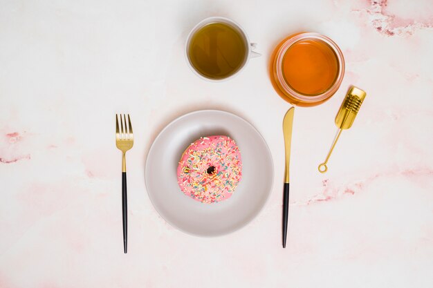 Tasse de thé vert; miel et beignet rose sur une plaque blanche avec une fourchette et un couteau à beurre sur fond blanc