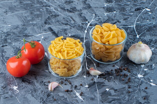 Tasses en verre de pâtes non cuites avec deux tomates fraîches et de l'ail sur une surface en marbre.